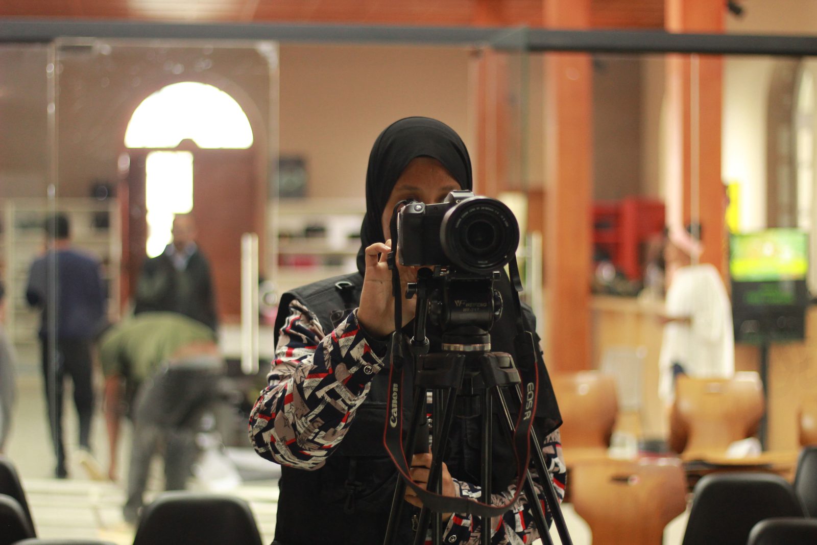 A camera operator frames a shot on a tripod-mounted camera in a busy hall.