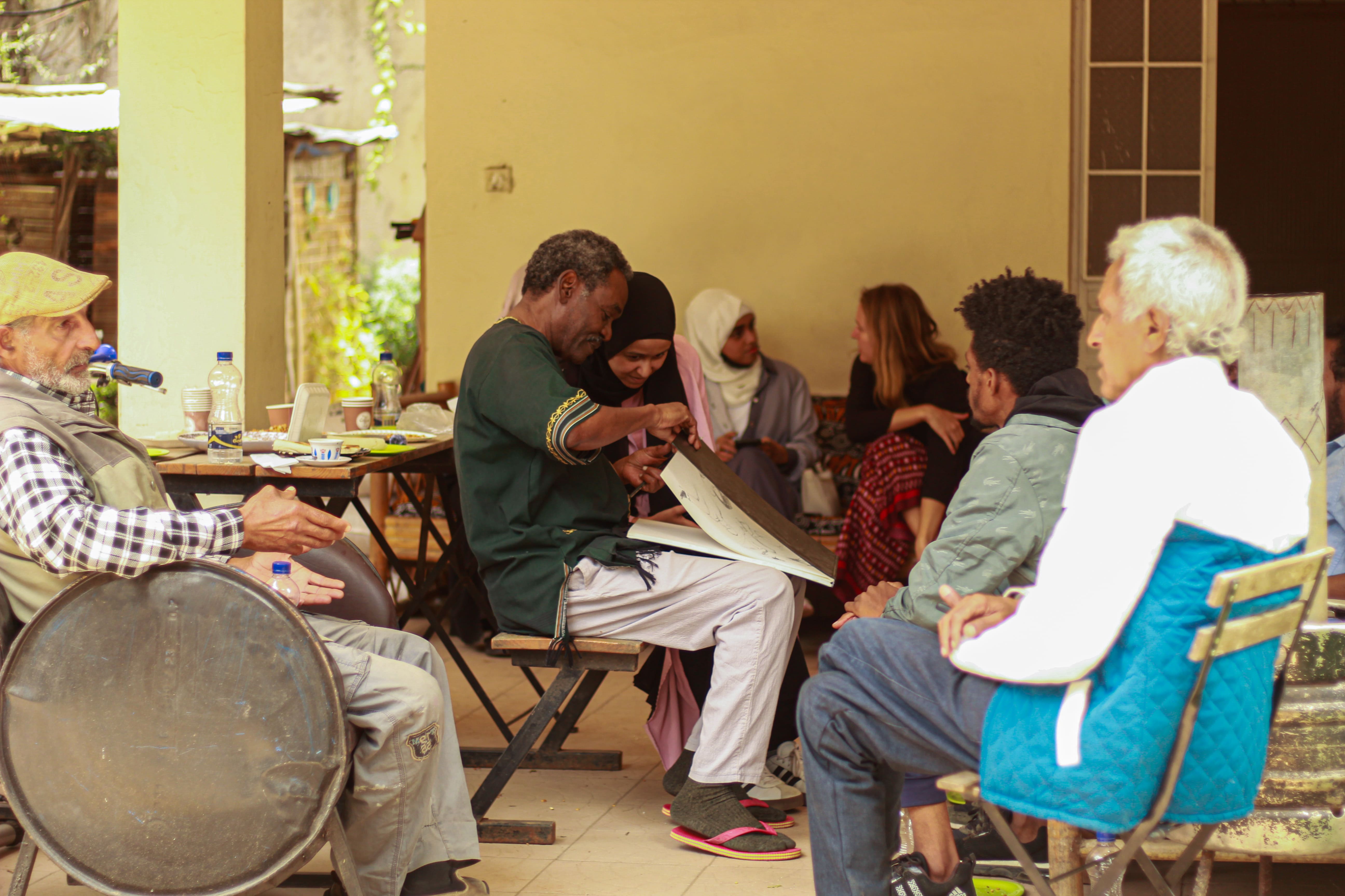 A man shows a sketchbook to the people seated around him at a community gathering on a veranda.